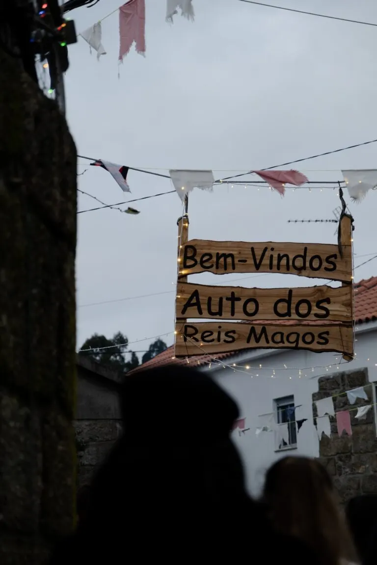 Placa de madeira «Bem-vindos Auto dos Reis Magos» suspensa sobre uma rua de Figueira, com bandeirolas e luzes