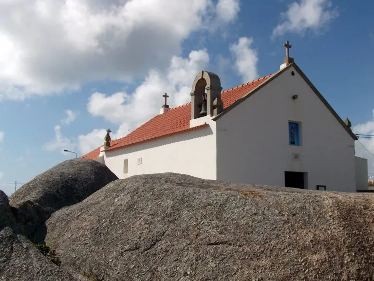 Capela de Santo André em Aver-o-Mar vista por trás de grandes penedos de granito, com o campanário e o telhado de telha