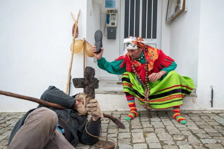 A Galdrapa, de saia mirandesa e lenços coloridos, ergue um sapato sobre o Velho caído no chão, junto à estaca com chouriça e bexigas
