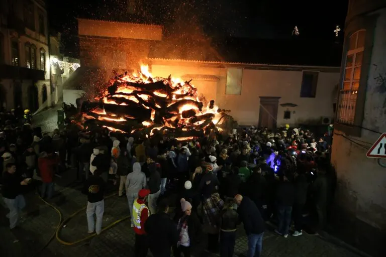 Madeiro aceso no adro da igreja, com faúlhas no ar e o público na rua à noite, em Penamacor