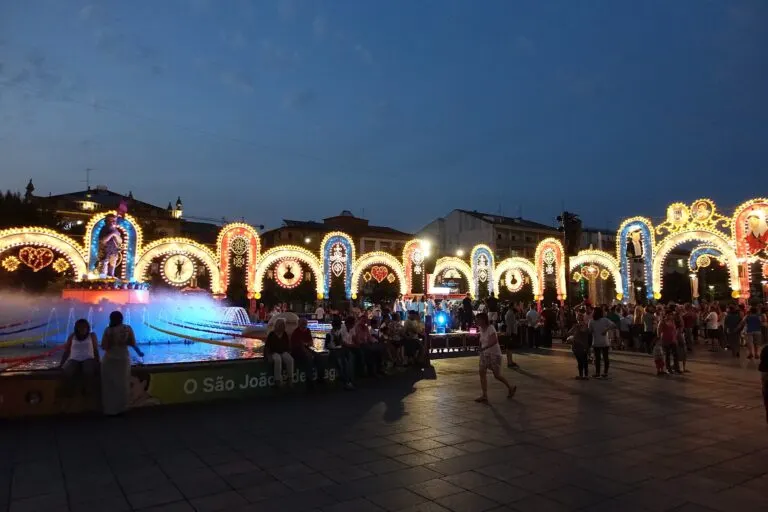 Fonte com a imagem de São João e arcos iluminados ao anoitecer, com gente a passear durante as festas de Braga